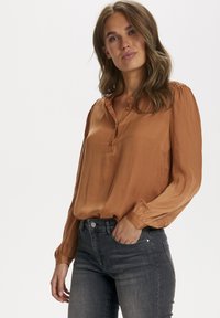 Blusa laranja queimada com um decote abotoado e uma textura de pontos subtil; mangas longas com punhos franzidos, combinada com jeans cinza escuro.