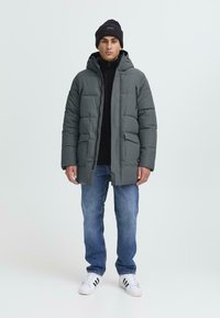 Jachetă puf în gri închis, cu glugă, având închidere cu fermoar, două buzunare frontale și interior din fleece, purtată peste un top negru. Blugi din denim și adidași albi.