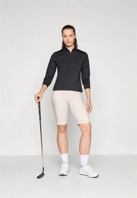 Noor naine mustas pika varrukaga topis ja beežides lühikestes pükstes seisab, hoides golfi putterit minimalisti halli tausta ees.