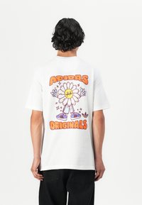 White cotton t-shirt featuring a colorful graphic of a smiling flower character with the text "ADIDAS ORIGINALS" prominently displayed.