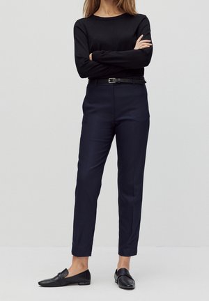 Vrouw staand met armen over elkaar, dragend een zwarte top met lange mouwen, marineblauwe op maat gemaakte broek met zwarte riem en zwarte leren loafers tegen een effen achtergrond.