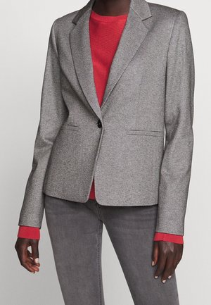 Femme portant un blazer gris sur un pull rouge avec un pantalon gris, debout devant un fond clair uni.