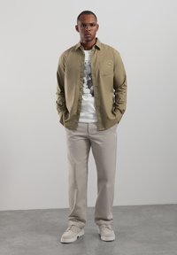 Chemise boutonnée couleur olive clair portée sur un t-shirt blanc à motif, associée à un pantalon beige et des baskets claires, sur un fond neutre.