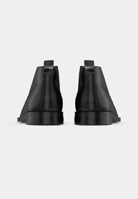 Μαύρες δερμάτινες μποτίνες μέχρι τον αστράγαλο με λεία υφή, στρογγυλή μύτη, κοντό φαρδύ τακούνι και χερούλια στο πίσω μέρος για εύκολη φόρμα.