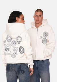 Cremefarvet hoodie med lynlås, der har forskellige sorte grafiske patches på ryggen og brystet. Akkompagneret af blå jeans med broderede bogstaver.