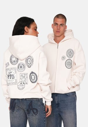 Creme kleurige hoodie met rits, voorzien van verschillende zwarte grafische patches op de achterkant en borst. Gecombineerd met blauwe spijkerbroek met geborduurde letters.