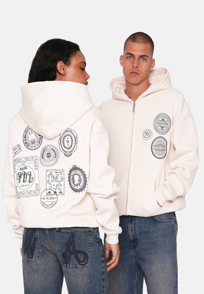 Cremefarvet hoodie med lynlås, der har forskellige sorte grafiske patches på ryggen og brystet. Akkompagneret af blå jeans med broderede bogstaver.