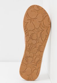 Roxy sandal sula i tan gummi med blommönster, som har präglade logotyper och texturerad yta för grepp. Konturerad design, platt form.