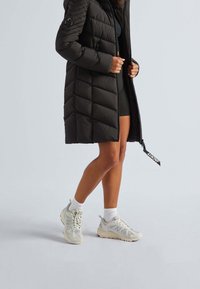 Schwarzer Steppmantel mit Chevron-Steppung, Reißverschluss vorne und Logo-Akzent. Getragen über schwarzen Shorts und hellgrauen New Balance Sneakern mit weißen Socken.