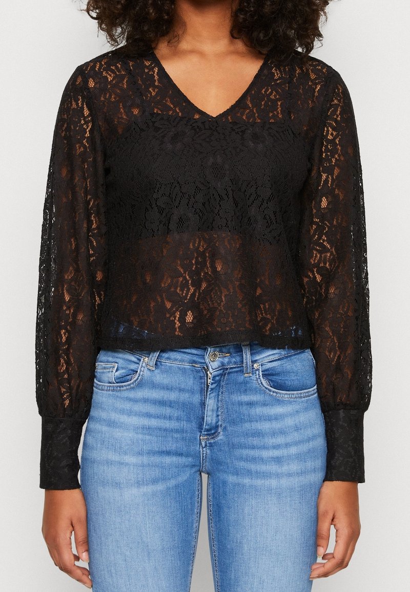 Černá krajková blůza s dlouhými rukávy, výstřihem do V a texturovaným květinovým vzorem, nošená přes světle modré džíny. Má volný střih.