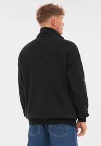 Pullover in pile nero con colletto alto, polsini a coste e vestibilità comoda, abbinato a jeans di denim blu. Tessuto morbido e design minimale.