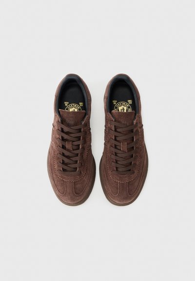 Brown suede sneakers with a low profile, featuring round laces, a textured toe cap, and a circular logo on the insole.