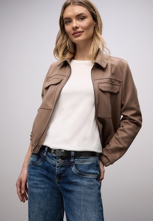 Młoda kobieta w beżowej kurtce na białej koszuli, w niebieskich dżinsach i czarnym pasku, stojąca na jednolitym tle.