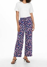 Kwiatowe spodnie w kolorze granatowym z różowymi, fioletowymi i zielonymi wzorami. Lekka tkanina, szerokie nogawki, zestawione z prostą białą koszulką i czarnymi sandałami.