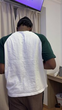 Ένα κοντομάνικο πουκάμισο με λευκό σώμα και πράσινα μανίκια, συνδυασμένο με καφέ παντελόνι με σχέδιο, που στέκεται κοντά σε μια ξύλινη επιφάνεια και μια τηλεόραση.