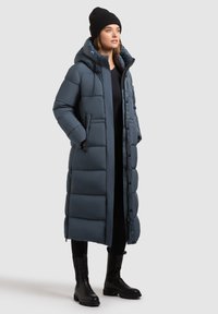 Lang, foret mørkeblå parka med hætte, der har lynlås og knapper foran, kombineret med sorte knælange støvler.