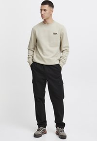 Beige sweatshirt med et lille patch, kombineret med sorte cargo-bukser med sidelommer og grå vandresko. Glat tekstur, afslappet pasform.