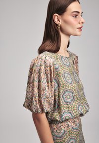 Blusa a fantasia con maniche a sbuffo, caratterizzata da disegni floreali e geometrici in tonalità di verde, rosa e blu, con una texture di tessuto liscia.