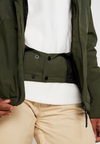 Haut en polaire blanc sous une veste vert olive avec rabat tempête à boutons-pression et pantalon beige sur une personne debout.