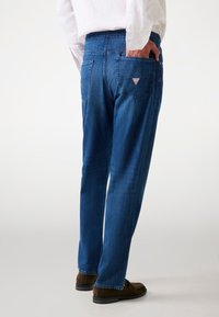Blue denim jeans with a relaxed fit, featuring back pockets, a logo patch on the right pocket, and a tapered leg design.