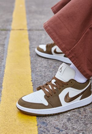 Brązowe i białe buty sportowe Nike Air Jordan z brązowymi sznurówkami, noszone z białymi skarpetkami i brązowymi spodniami na asfalcie w pobliżu żółtej linii.