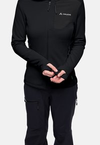 Giacca nera con zip e fori per il pollice, cappuccio regolabile e tasche laterali. Accentuata da un logo bianco sul petto. Indossata con pantaloni scuri.