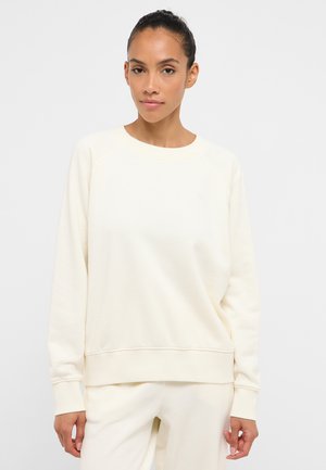 Woman with dark hair tied back wearing a cream-colored long-sleeve sweatshirt and matching pants, standing against a plain background.