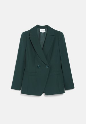 Kvinde iført en mørkegrøn buksedragt med en blomster-silke-bluse, sort bælte, sorte høje hæle og metallisk manchetarmbånd, stående med den ene hånd i lommen.