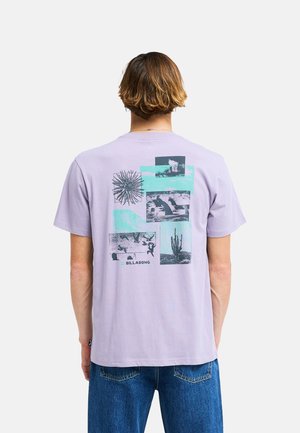 Homme aux cheveux courts et bruns portant un t-shirt Billabong violet clair avec des images graphiques bleues et un jean bleu, vu de dos.