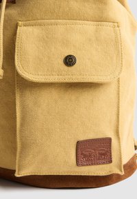 Geel canvas rugzak met voorvak met knoopsluiting en een bruine leren merkembleem van Levi Strauss & Co. nabij de onderrand.