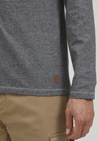 Grå langermet skjorte med teksturert strikk, som har et lite brunt lærlogo-patch nær sømmen og kontrastsøm langs kantene.
