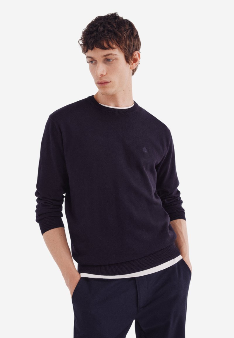Marineblauer Rundhalsausschnitt-Pullover mit gerippten Bündchen und Saum, aus einem glatten Stoff gefertigt, über einem weißen Hemd getragen, kombiniert mit dunklen Hosen.