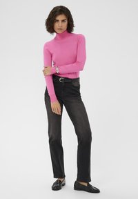 Pull à col roulé rose, coupe ajustée, associé à un jean en denim foncé. Le mannequin porte des mocassins noirs, un bracelet en argent, sur un fond uni.