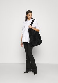 Saco de pele preta em cotovelo com design em camadas, apresentando uma alça resistente. Modelo vestindo uma camisa branca de botões com bolinhas e calças pretas.