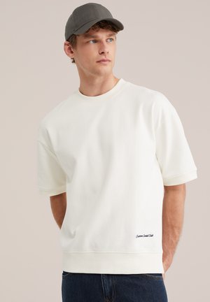 Jeune homme aux yeux bleus portant une casquette grise, un sweat-shirt blanc oversize à manches courtes et un jean foncé, regardant sur le côté sur un fond neutre.