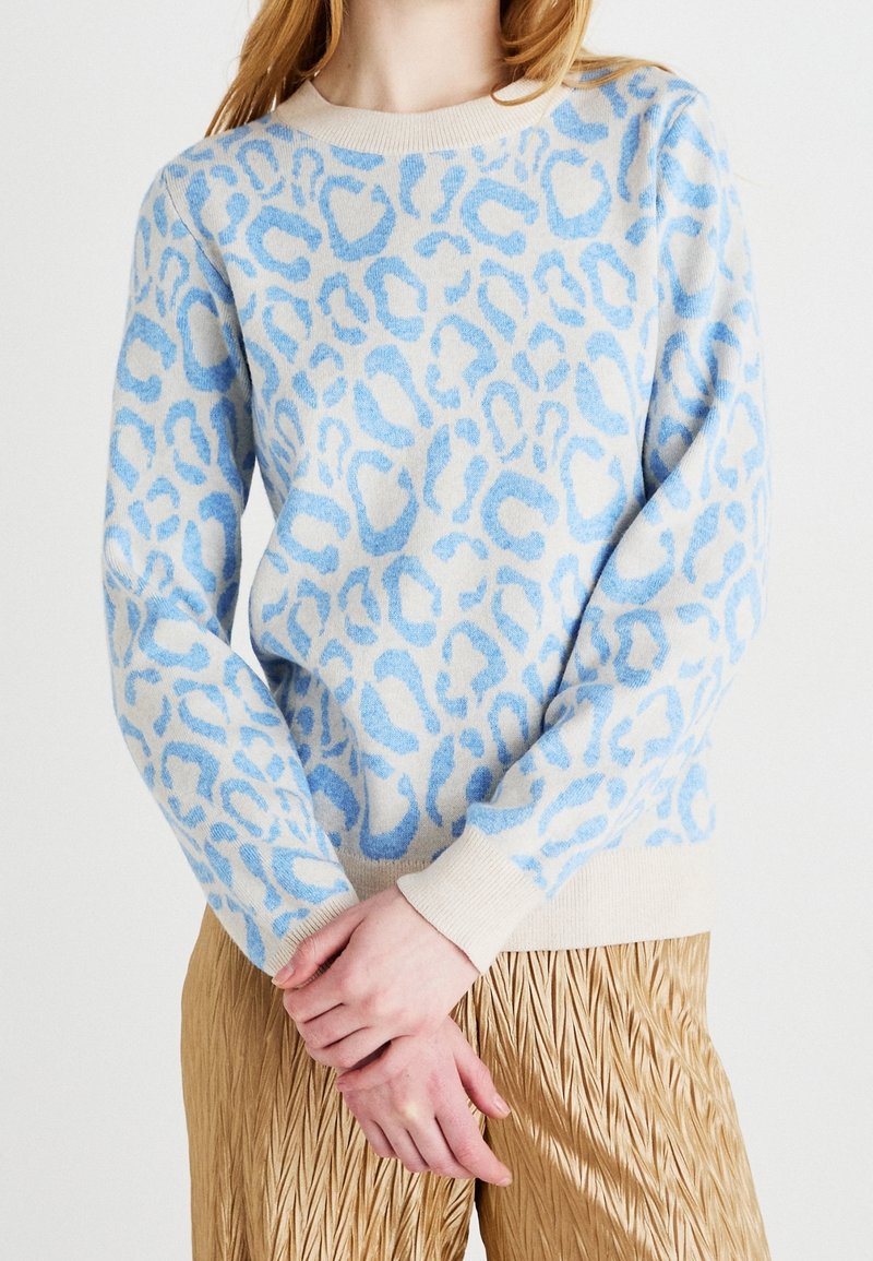 Femme portant un pull beige à imprimé léopard bleu clair et un pantalon plissé doré, les mains doucement croisées à la taille.