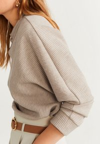 Beige Rippstrickpullover mit loser Passform, hohem Kragen und umgeschlagenen Bündchen. Kombiniert mit hochgeschnittenen beigen Hosen und einem braunen Gürtel.