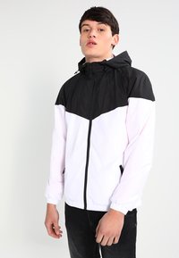 Veste coupe-vent en coloris bloqués avec une partie supérieure noire et une partie inférieure blanche. Comprend une fermeture éclair, une capuche réglable et des poignets élastiques.