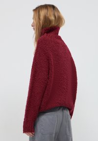 Frau mit langen Haaren, die einen strukturierten burgunderfarbenen Pullover mit hohem Kragen und lockeren grauen Hosen trägt, im Profil vor einem neutralen Hintergrund stehend.