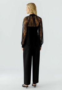 Schwarze Spitzenbluse mit langen Ärmeln über einem einfarbigen schwarzen Oberteil. Sie hat eine durchsichtige Textur und florale Muster. Kombiniert mit weit geschnittenen schwarzen Hosen.