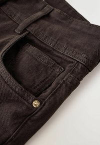 Gros plan sur un jean en denim marron foncé montrant la poche avant, la poche à monnaie, la passants de ceinture et un détail de rivet en métal sur un fond clair.