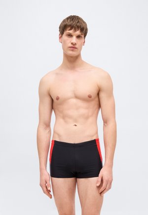 Bărbat tânăr cu păr scurt și brunet, purtând slip de baie negru cu panouri laterale roșii, stând în picioare pe un fundal simplu, deschis la culoare.