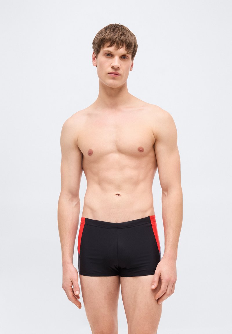 Νεαρός άνδρας με κοντά καστανά μαλλιά, που φοράει μαύρο μαγιό με κόκκινες πλαϊνές λωρίδες, στέκεται μπροστά σε ένα απλό φωτεινό φόντο.