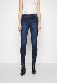 Tmavě modré skinny džíny s vysokým pasem, se předními kapsami a metalickým knoflíkem na zapínání. Kombinované se světlým topem a černými kotníkovými botami.