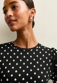 Zwarte textuurtop met wit diamanten patroon, met een ronde halslijn en geplooide mouwen. Gouden druppeloorbellen accentueren de look.