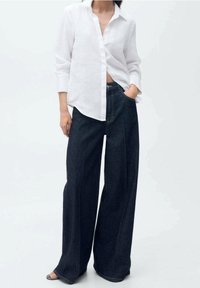 Chemise en lin blanc à boutons avec manches longues, associée à un jean en denim foncé à jambes larges. La tenue présente une coupe décontractée et un design casual.