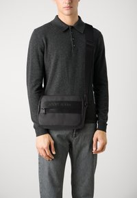 Borsa a tracolla nera con una texture liscia, dotata di una tasca con zip, logo impresso e tracolla regolabile. Indossata sopra un maglione scuro.