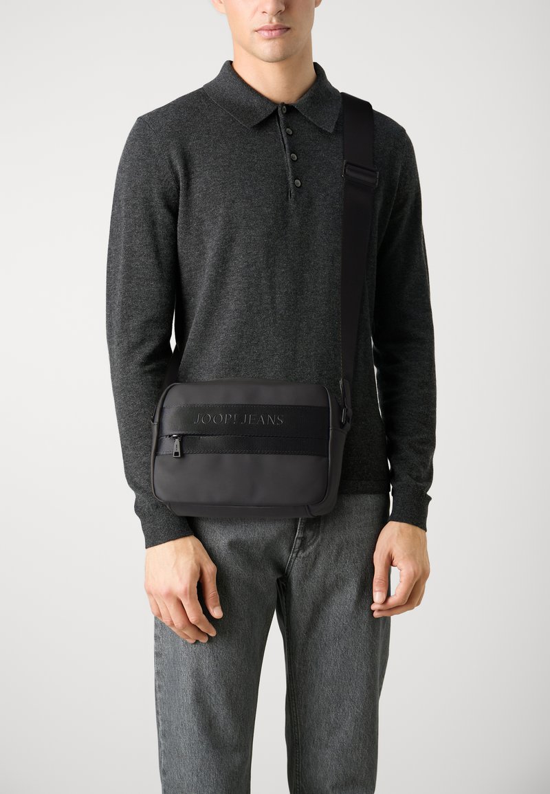 Borsa a tracolla nera con una texture liscia, dotata di una tasca con zip, logo impresso e tracolla regolabile. Indossata sopra un maglione scuro.