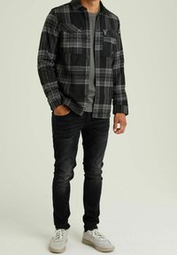 Camicia di flanella a scacchi nera e grigia con bottoni frontali, tasche sul petto e una maglietta girocollo grigia sotto. Abbinata a jeans scuri e sneakers chiare.