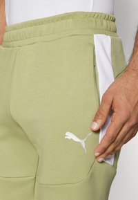 Calças de fato de treino verde claro com uma textura suave, detalhes brancos nas laterais e um logo branco da Puma na parte inferior da perna.